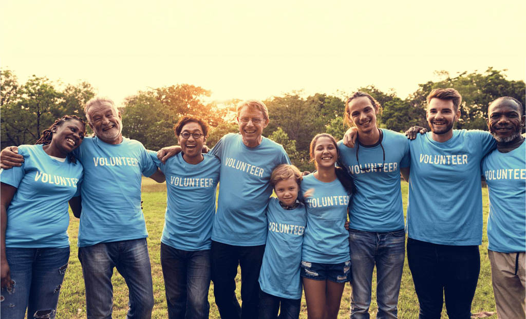 Chancen för Vereine En Grupp di Mensch, wat sich zusammenfuhren und en T-Shirt hat, wat das Wort "VOLUNTEER" aufweist. Sie stehn in en offener Gelände mit en Sonnenuntergang-Szene im Hintergrund.